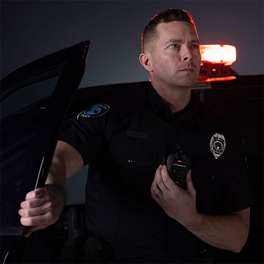A uniformed male police officer stands next to a patrol car with red emergency lights on with the door open, holding the frame with one hand and resting his other hand on a chest-mounted Motorola Solutions body camera while looking off to the side with a serious expression.