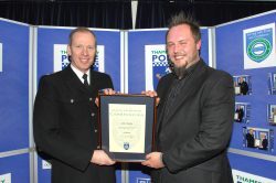 The image shows Carl, a man with a beard, receiving a commendation for his work in the Thames Valley Police control room. He is smiling and holding a framed certificate while standing next to a police officer in uniform.