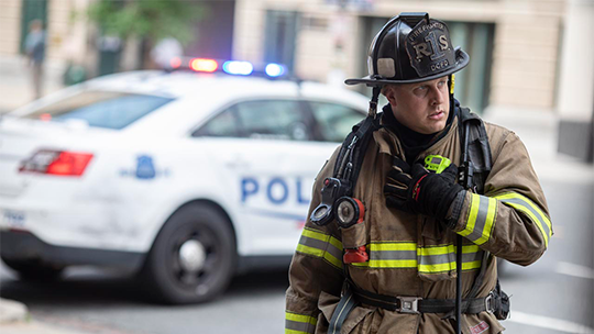 Fireman talking into a radio with a police car in the background